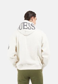 Sudadera crema con el logo "GUESS" en gris en la capucha. Presenta puños acanalados, un corte relajado y un diseño liso en la parte posterior. Adecuada para uso diario.
