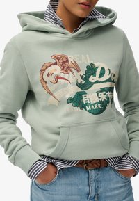 Persona che indossa una felpa leggera verde con ricami di uccelli e onde sopra una camicia a righe con colletto e jeans con cintura intrecciata.