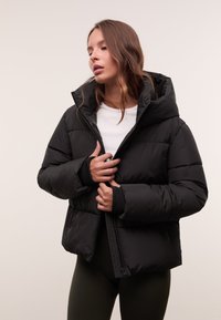 Eine Frau mit langen Haaren, die eine schwarze Pufferjacke, ein weißes Shirt und dunkle Hosen trägt. Sie hält den Reißverschluss der Jacke halb geschlossen vor einem schlichten Hintergrund.