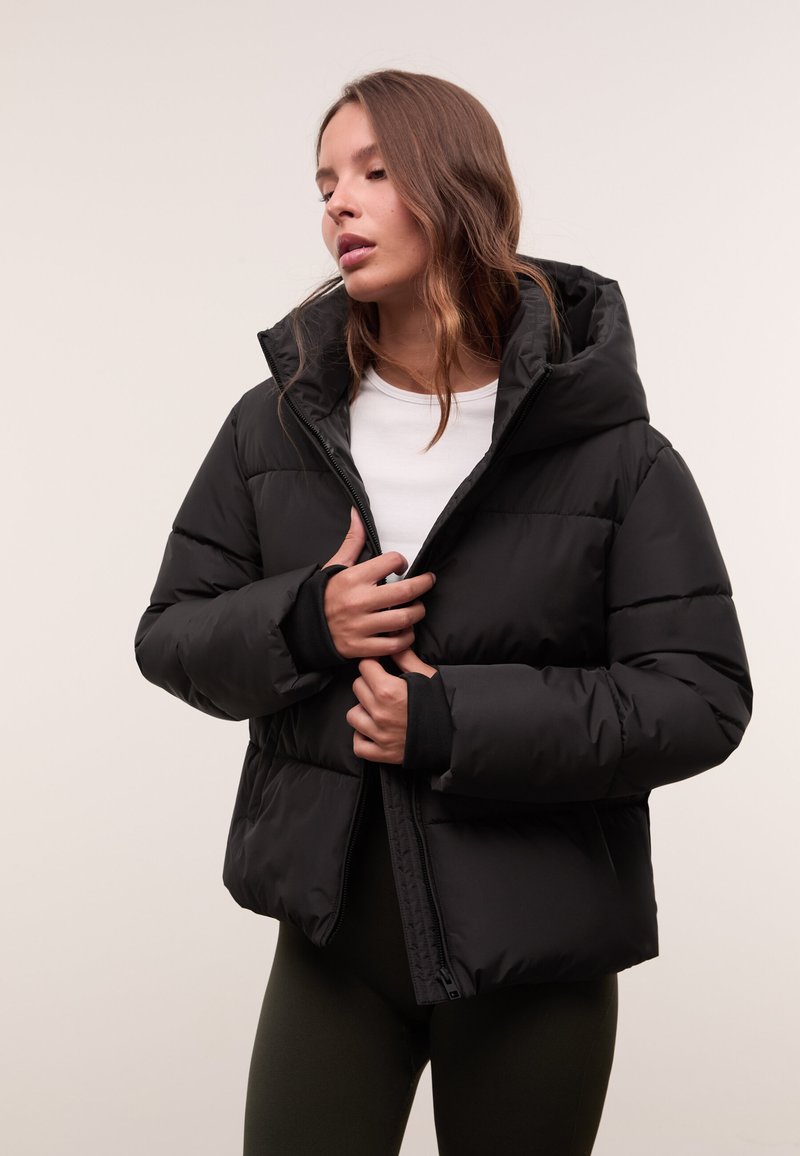 Eine Frau mit langen Haaren, die eine schwarze Pufferjacke, ein weißes Shirt und dunkle Hosen trägt. Sie hält den Reißverschluss der Jacke halb geschlossen vor einem schlichten Hintergrund.