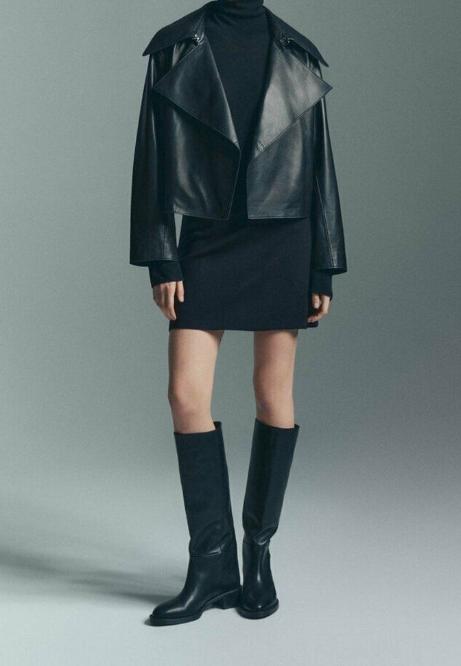 Veste courte en cuir noir à larges revers, associée à une robe noire cintrée et des bottes en cuir noir montantes jusqu'au genou. Textures lisses sur l'ensemble.