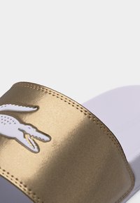 Κοντινή λήψη ενός χρυσού λουριού σανδαλιού τύπου σλάιντ με λευκό ανάγλυφο λογότυπο κροκόδειλου και λεπτομερές ράψιμο σε ανοιχτό φόντο.