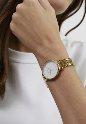 Montre en métal doré avec un cadran blanc rond, des aiguilles fines et un bracelet à maillons. Portée au poignet, avec une chemise blanche unie en arrière-plan.