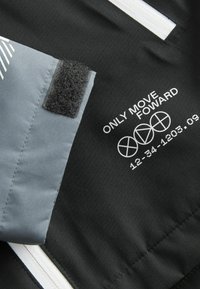 Sort stof med en struktureret grå lomme med velcro. Hvid tekst og symboler siger "BEVÆG KUN FREMAD" med tal nedenfor.