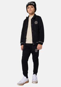 Haut à capuche noir avec des boutons et un logo, associé à un pantalon de jogging assorti. Des baskets blanches avec des accents gris complètent la tenue. Bonnet noir avec un logo en texte.