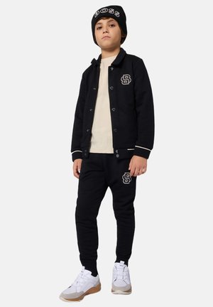 Felpa nera con bottoni e logo, abbinata a pantaloni della tuta coordinati. Sneakers bianche con accenti grigi completano l'outfit. Berretto nero con logo testuale.