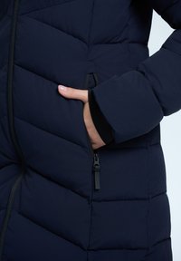 Námořnická puffer bunda s prošívanou texturou, zipovou kapsou a elastickými manžetami pro pevné uchycení. Měkká podšívka přidává na pohodlí.