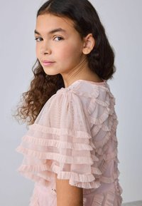 Hosszú, göndör hajú lány világos rózsaszín fodros ruhában, a vállán túlnézve, egyszerű háttér előtt.