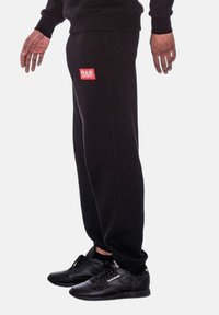 Schwarze Sweatpants aus weichem Stoff, mit einem roten Logopatch auf der Vorderseite. Lockere Passform mit seitlichen Taschen, kombiniert mit schwarzen Turnschuhen.