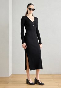 Black ribbed long-sleeve dress with a V-neck and button detail. Features a side slit and worn with black heeled shoes.