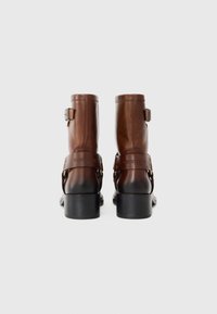Bottines en cuir marron avec une sangle à boucle, bout arrondi et talons en bloc noirs. Texture lisse, couture minimale.