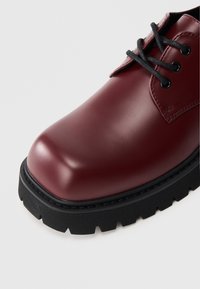 Bordó bőr cipő fényes felülettel, lekerekített orral, fekete fűzőkkel és vastag fekete gumitalppal, texturált mintázattal.