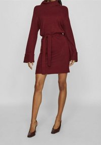 Robe tricotée bordeaux avec un col montant, une taille ceinturée et de larges poignets de manche. Le tissu semble doux et texturé. Chaussures à talons hauts marron.