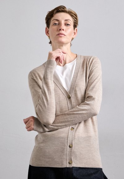 Beiger Cardigan aus weichem Material mit V-Ausschnitt, langen Ärmeln und sechs vorderen Knöpfen. Über einem weißen T-Shirt und dunklen Jeans getragen.