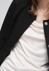 Blazer negro con un diseño entallado y detalles en metal plateado, llevado sobre una blusa drapeada brillante de color crema.