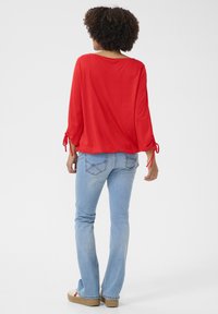 Red, lightweight top with three-quarter sleeves featuring adjustable ties at the cuffs. Paired with light blue flared jeans.