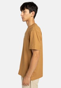 Camiseta de algodón marrón con cuello redondo, mangas cortas y costuras laterales. Presenta un pequeño diseño en el pecho, dobladillo plano y un corte relajado.