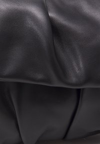 Musta nahkamateriaali, jossa on teksturoitu pinta, pehmeät, poimutetut taitteet ja sileä pinta. Minimalistinen muotoilu ilman näkyvää metalliosaa.