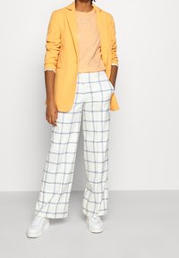 Osoba v žlutém saku, broskvovém tričku Nike, bílých širokých kalhotách s modrým kostkovaným vzorem a bílých teniskách, stojící s rukama v kapsách.