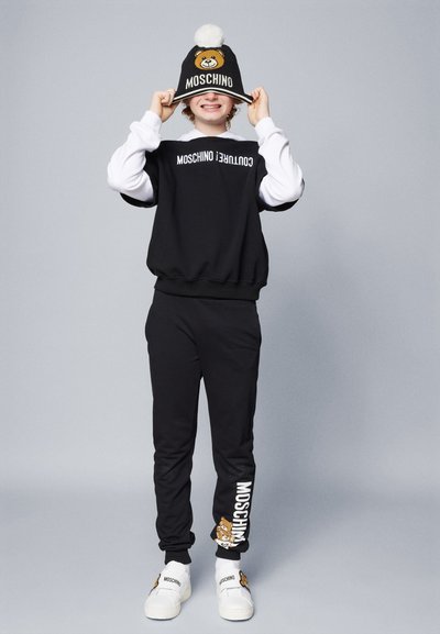 Μαύρη φόρμα με μανίκια αντίθετης λευκής απόχρωσης· διαθέτει λογότυπο Moschino. Μαύρο σκουφάκι με σχέδιο αρκούδας και λευκή φούντα. Λευκά slip-ons.