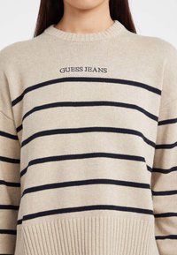 Beidzītas adījuma džemperis ar melnām horizontālām svītrām un uz krūtīm izšūtu uzrakstu "GUESS JEANS", ko valkā persona ar gariem tumšiem matiem.