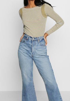 Mujer con camiseta de manga larga de rayas oliva y blancas metida en vaqueros de pierna ancha azul claro de talle alto, de pie con las manos en las caderas.