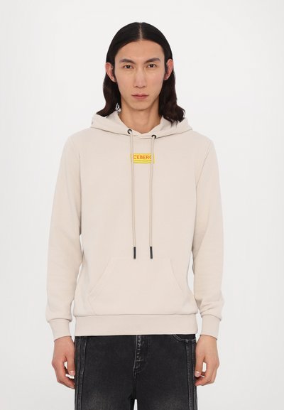Haut à capuche beige avec poche kangourou à l'avant, cordons de serrage réglables et un patch rectangulaire jaune. Le tissu semble lisse et légèrement épais.