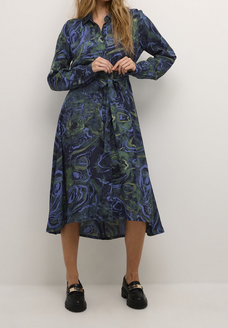 Femme portant une robe à manches longues boutonnée avec ceinture nouée à la taille, aux tourbillons abstraits bleus et verts, associée à des mocassins noirs avec détails dorés.