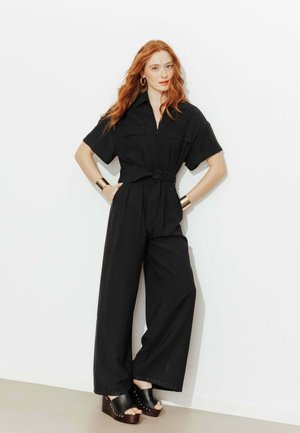 Femme rousse portant une combinaison noire ceinturée, des boucles d'oreilles créoles dorées, des bracelets dorés et des sandales noires compensées, debout contre un mur blanc.