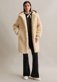 Crème faux fur jas met een oversized pasvorm, capuchon en zijzakken; gedragen over een donkere top en gecombineerd met zwarte flared pants en witte sneakers.