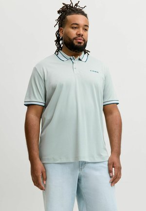 Muškarac s pletenicama, odjeven u svijetloplavu polo majicu s tamnim obrubom i svijetloplave traperice, stoji ispred jednobojne pozadine.