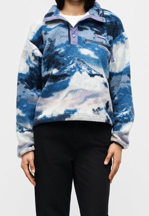 Person iført en blå, hvid og lilla mønstret fleece-pullover med en lynlåslomme på brystet, kombineret med sorte bukser, stående mod en ensfarvet baggrund.