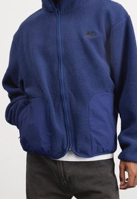 Blå fleecejacka med hög krage, dragkedjestängning och sidofickor; har en slät tygdetalj på fickorna och en Nike-logotyp.