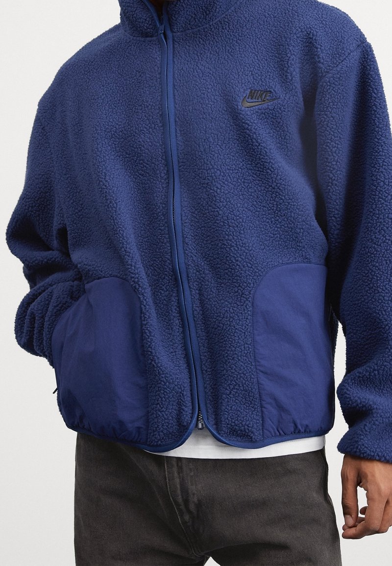 Blå fleecejacka med hög krage, dragkedjestängning och sidofickor; har en slät tygdetalj på fickorna och en Nike-logotyp.