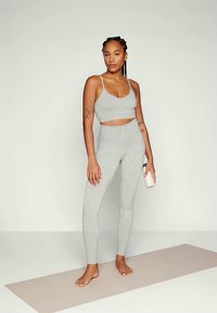 Grijze sportieve crop top en hoge taille leggings van rekbaar materiaal. Het model houdt een waterfles vast en staat op een yogamat.