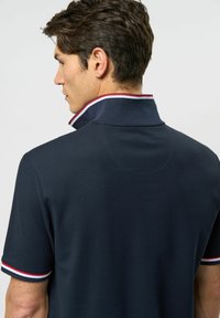 Laivastonsininen polo-paita, jossa on korkea kaulus ja punaisia sekä valkoisia raitoja kauluksessa ja hihansuissa. Suunnittelu on sileäpintaista ja istuvaa.