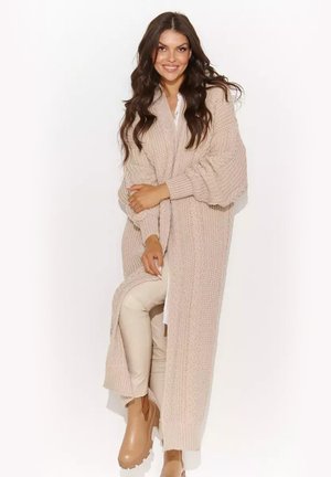 Kobieta o długich ciemnych włosach, ubrana w beżowy gruby dzianinowy długi kardigan, jasne spodnie i brązowe botki na masywnej podeszwie, uśmiechająca się i pozująca.