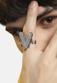 Sølvfarvet ring med en "V"-form, prydet med klare krystaller og grønne accenter, der har en struktureret overflade og metallisk finish.