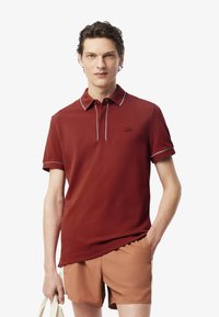 Röd polo skjorta med krage, vita detaljer och korta ärmar, kombinerad med beige shorts. Tyget verkar slätt och lätt.