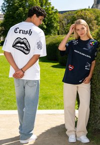Deux jeunes adultes se tiennent à l’extérieur ; l’un porte un t-shirt blanc « Crush » avec un motif de lèvres, l’autre un t-shirt noir avec des patchs et un pantalon beige.