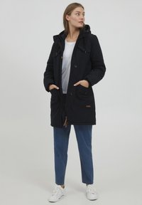 Marineblauwe gewatteerde parka met een capuchon, voorzien van knoopsluitingen, meerdere zakken en een trekkoord. Draagt over een wit shirt en blauwe jeans.