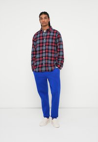 Rødt og marineblåt ternet flannel skjorte med knapper, parret med blå sweatpants. Lysfarvede sneakers fuldender outfittet. Hvid baggrund.
