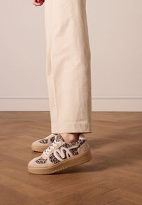 Leopardenmuster-Sneakers aus beigem Wildleder mit weißen Akzenten und einer strukturierten Gummisohle. Getragen mit creamfarbenen weiten Hosen auf einem Holzboden.