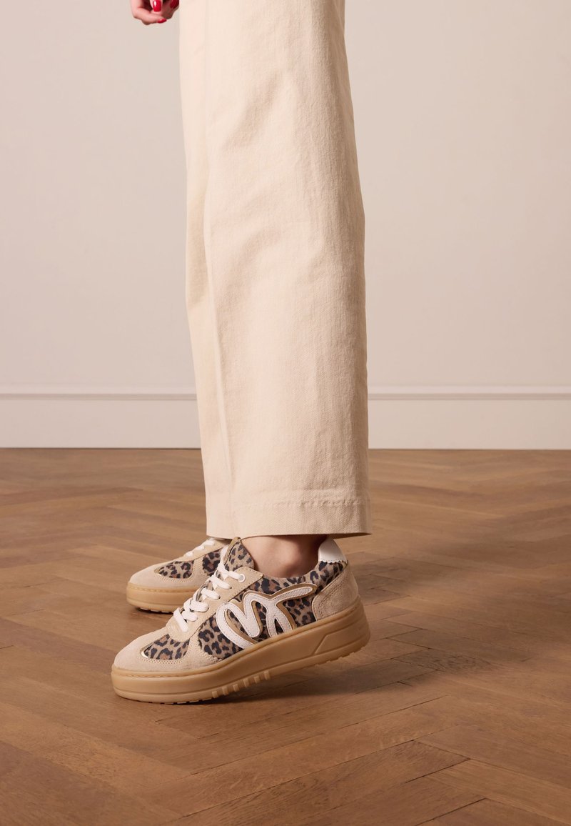 Leopardenmuster-Sneakers aus beigem Wildleder mit weißen Akzenten und einer strukturierten Gummisohle. Getragen mit creamfarbenen weiten Hosen auf einem Holzboden.