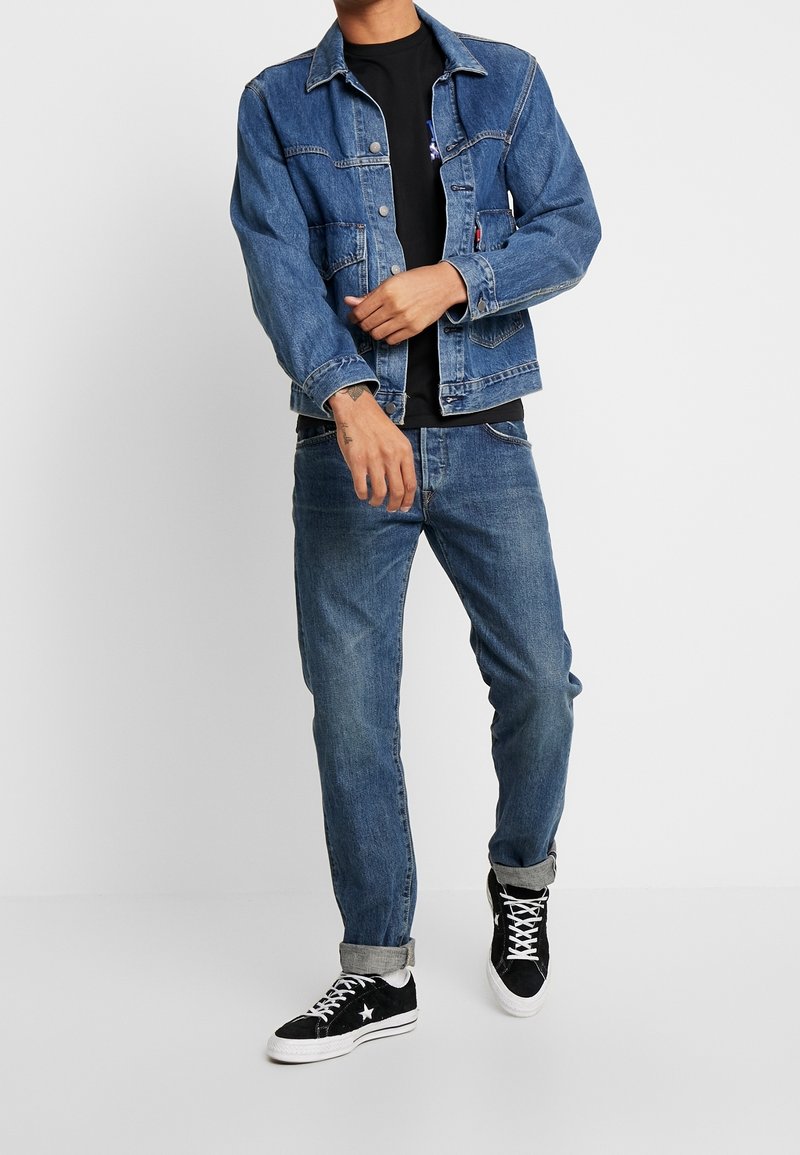 Homme portant une veste en jean bleu et un jean, un t-shirt noir, et des baskets noires avec des lacets blancs, posant avec les mains près de la taille.