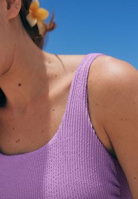 Fioletowy teksturowany strój kąpielowy z szerokim paskiem, prezentujący prążkowany wzór i gładką tkaninę, na tle oświetlonej słońcem skóry i niebieskiego nieba.