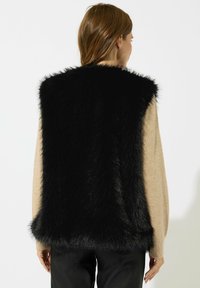 Svart fuskpälsväst med en fluffig textur och ärmlös design. Buren över en tanfärgad tröja, har en ren, rak fåll.