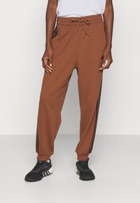 Person wearing brown Adidas sweatpants with black stripes down the sides and black athletic shoes, standing against a plain background.