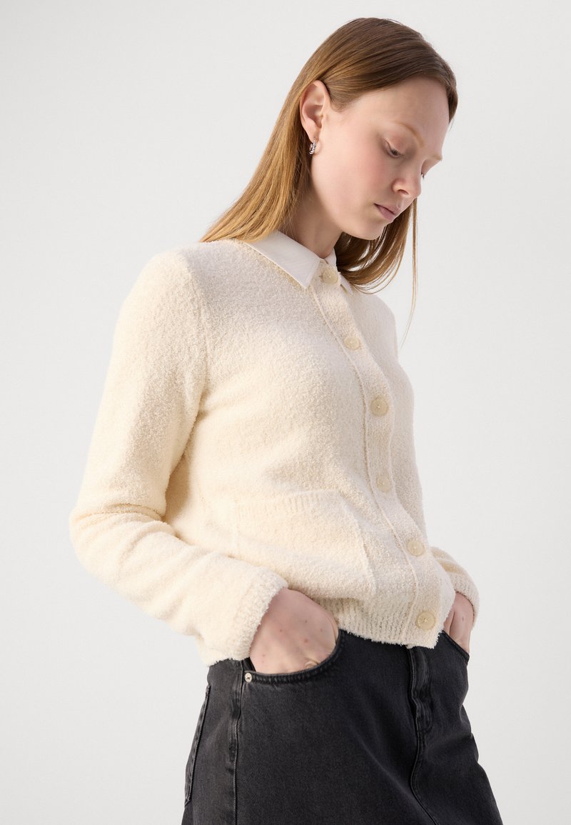 Cremefarbener, fuzziger Cardigan mit Knopfverschluss, Polokragen und zwei Taschen. Über einem dunkelblauen Jeansrock getragen.