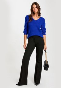 Maglione lavorato a maglia blu con scollo a V e maniche arrotolate, abbinato a pantaloni neri a vita alta e a campana. La modella tiene una borsa nera con tracolla a catena.